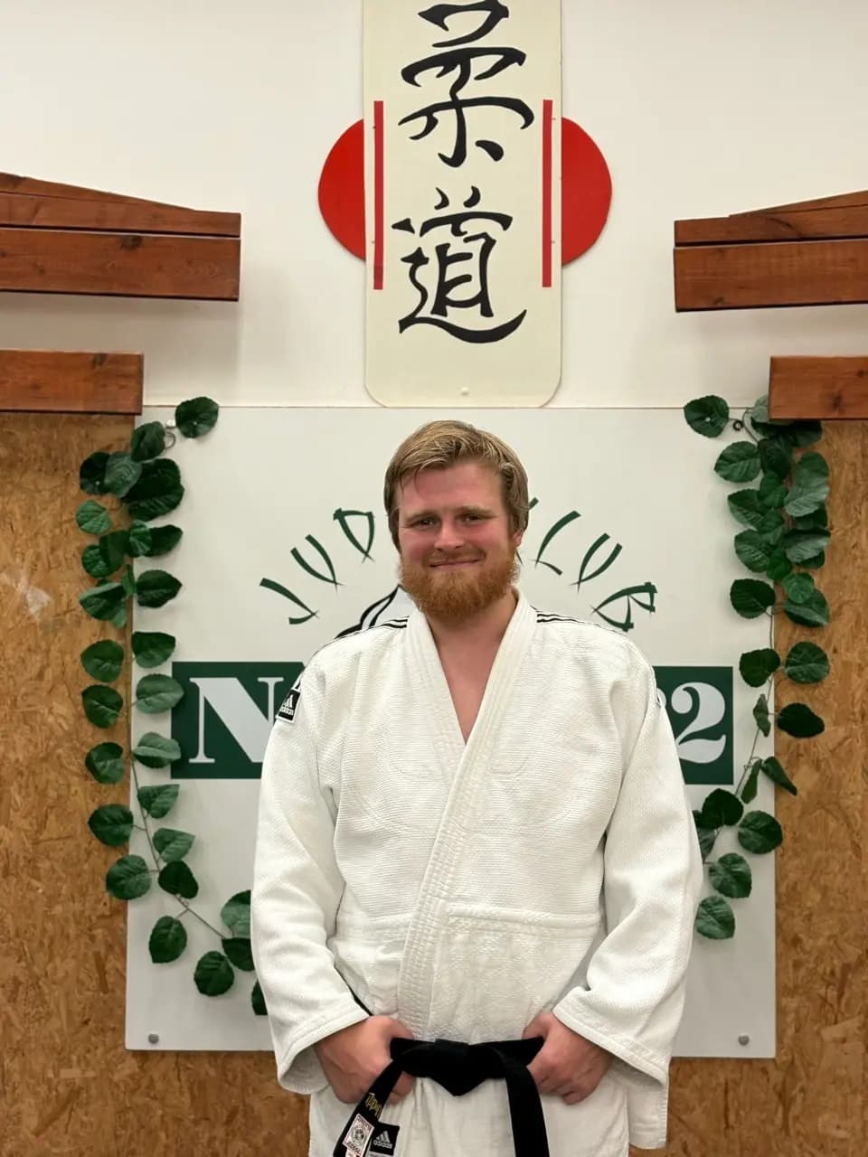 Trenér Ing. Petr Košut v bílém judogi s černým pásem před klubovou tabulí a kaligrafií 柔道