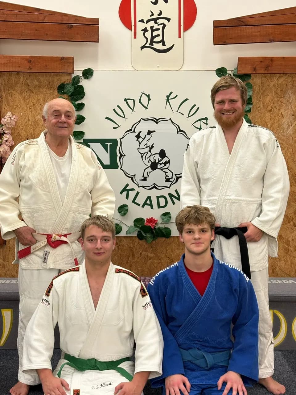 Čtyři judisté oddílu v judogi v dojo, za nimi na stěně zelené logo Judo klub N Kladno